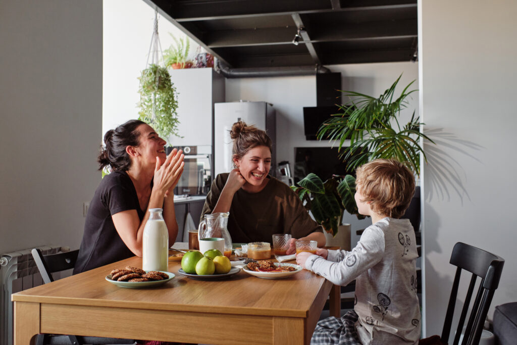 Happy lesbian couple laughing during breakfast at table while their little son talking funny jokes